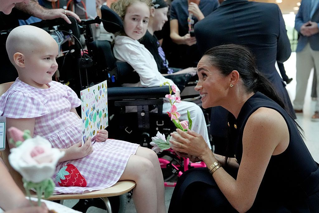 The Duchess of Sussex receives flowers and a card from a young patient on a visit to the Royal Children's Hospital in Melbourne. The Duchess and patient are smiling at one another.