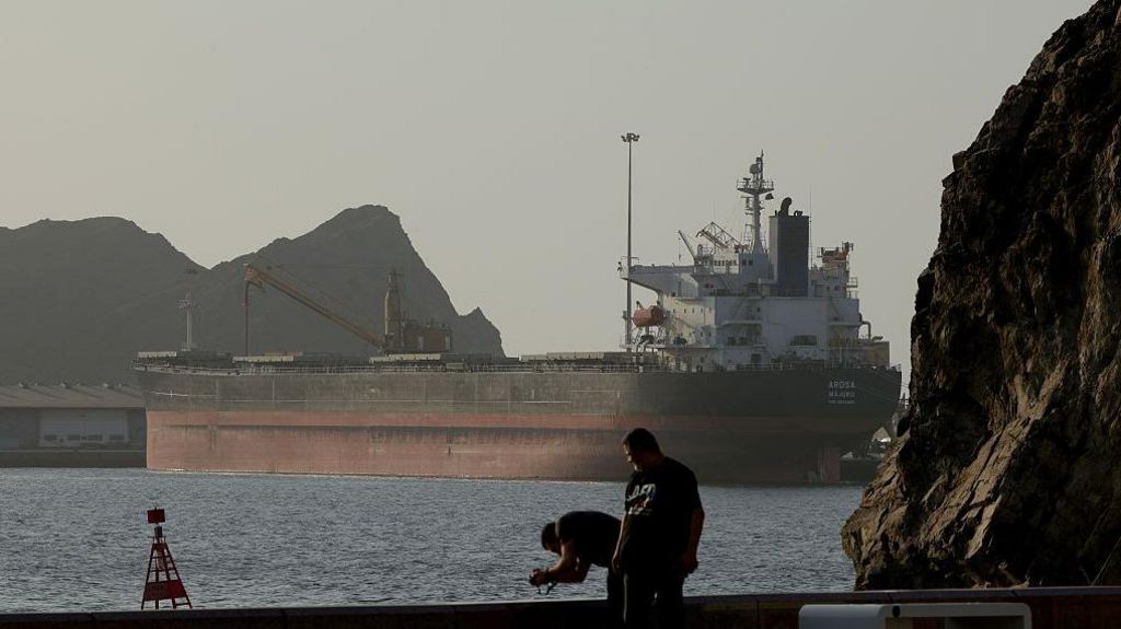 An oil tanker stands anchored between two rocky shores on a sunny evening. The image shows the Strait of Hormuz on 17 June
