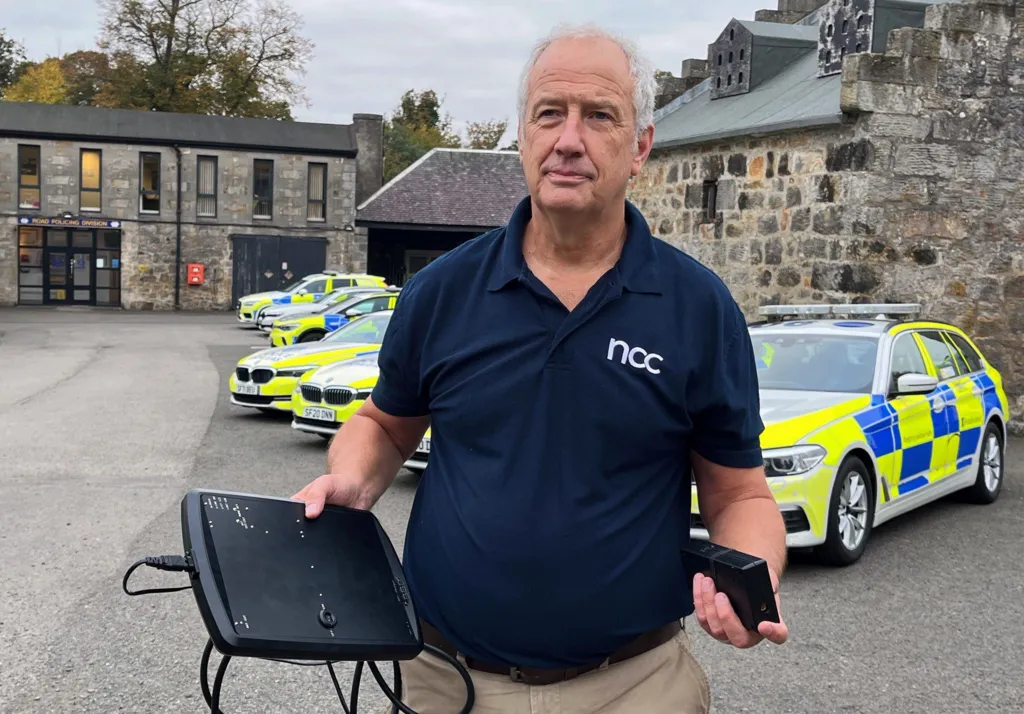 A grey haired man in a dark short sleeved top holding two black electronic boxes. In the background are a number of police cars