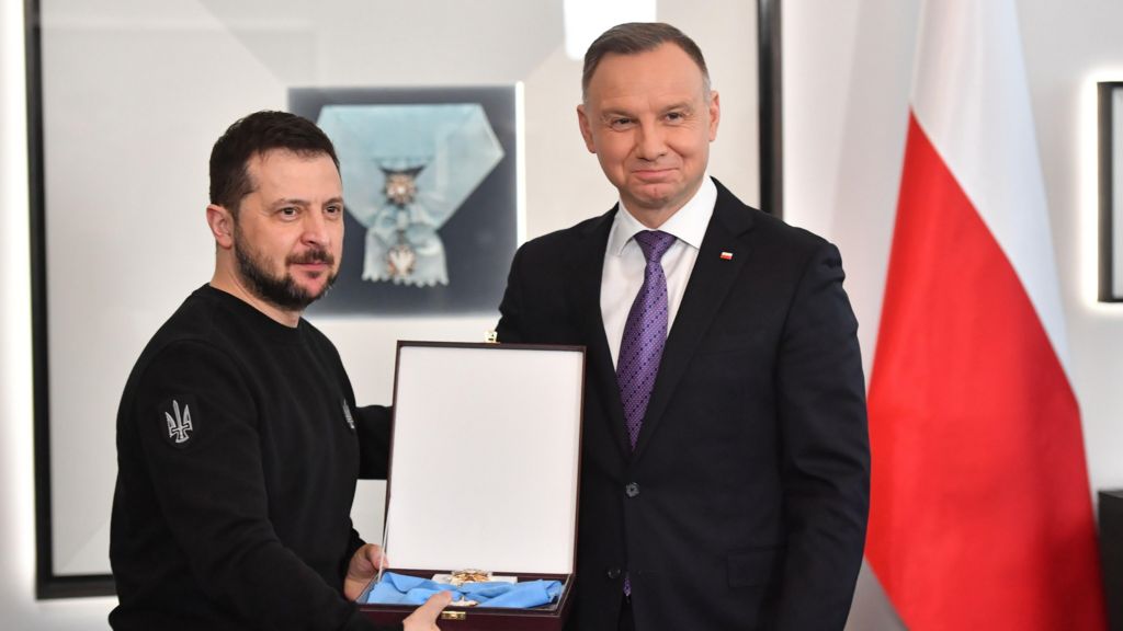 Polish President Andrzej Duda (R), wearing a suit and tie, awards Ukrainian President Volodymyr Zelensky, wearing a black-long sleeve shirt, with the Order of the White Eagle during his visit in Warsaw, Poland. The medal is lying in an opened box, which Zelensky is holding.
