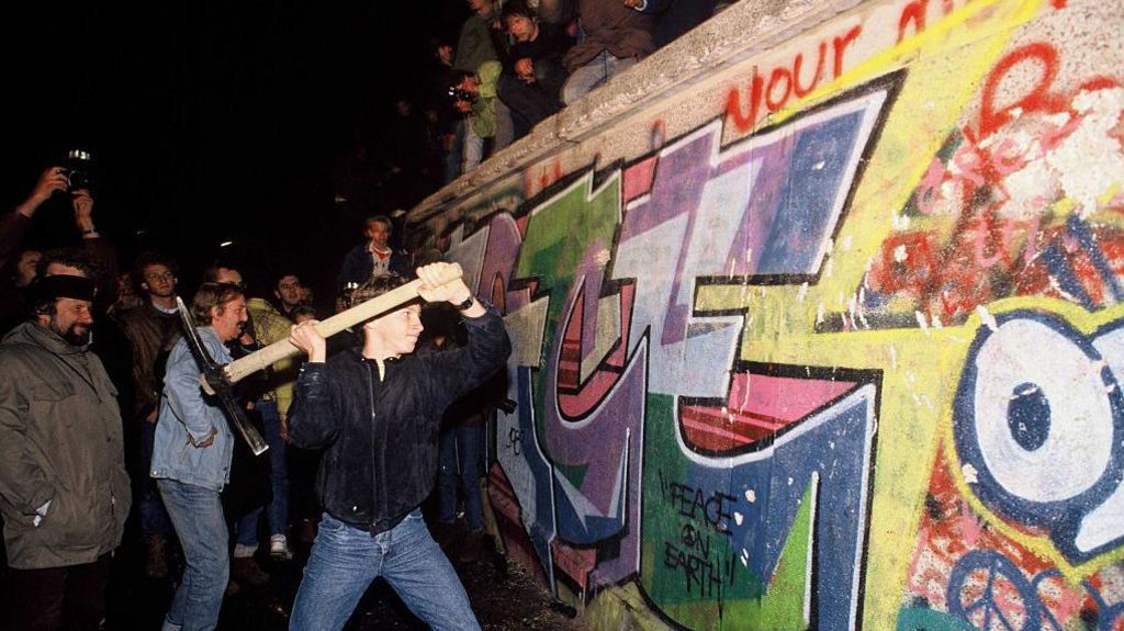 A man uses a pickaxe to smash the Berlin wall, surrounded by an enthusiastic crowd.
