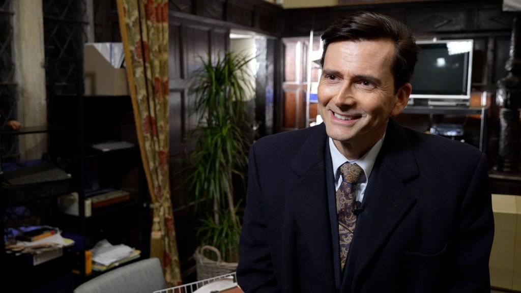 David Tennant smiles, looking off camera. He's wearing a black coat with shirt and tie. In the background is a television and a dark-wood panelled wall, with a big curtain that has green and red flowers.