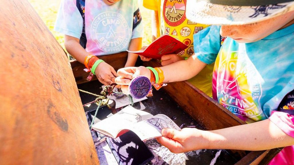 A child holds a stamp over a book in which he has just put the stamp's impression on using purple dye. The child wears a multi-coloured T-shirt and has a hat obscuring most of his face