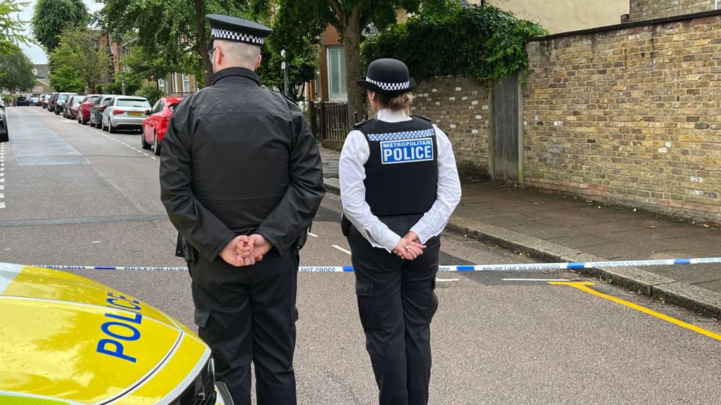 Two Metropolitan Police officers standing with their backs to the camera behind police cordon tape on a residential street.