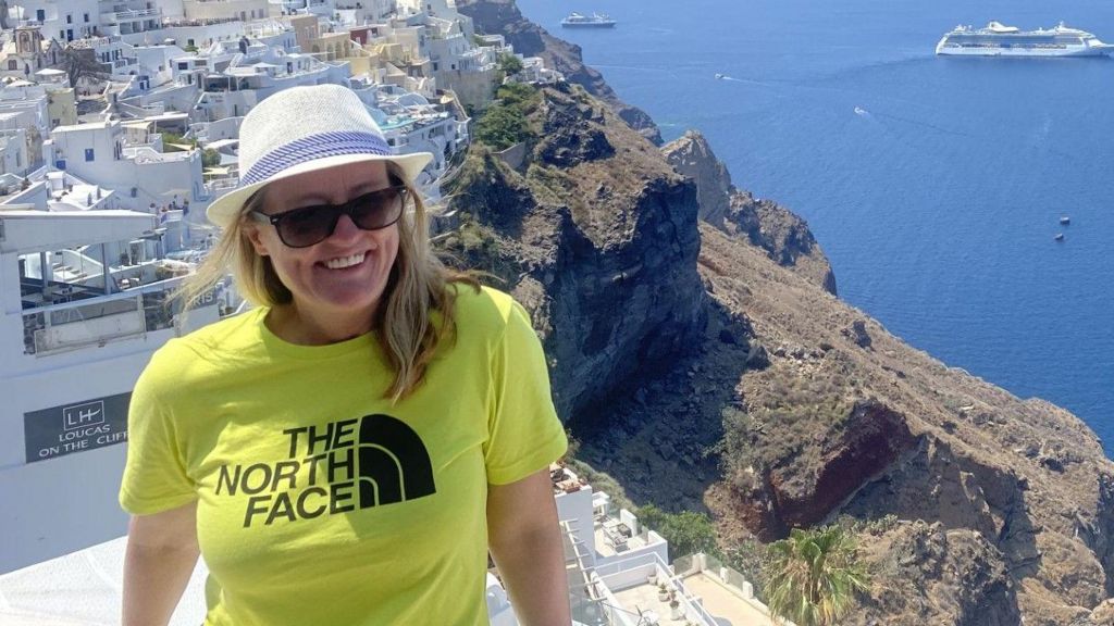 Ellen Mulvey on holiday abroad with long blond hair wearing black sunglasses and a white and blue sun hat and lime green t-shirt with the words 'The North Face' on it. She is standing on a property on a hill by the sea. She is smiling.