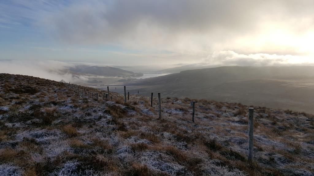 A frosty scene with a wooden fence, a peaty landscape and a reservoir in the far distance 