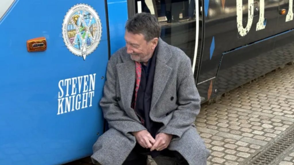 Steven Knight, wearing black suit and grey coat, is crouching by the side of a blue section of tram bearing his name. There is cobbled street beneath the tram and a drainage grid running alongside.