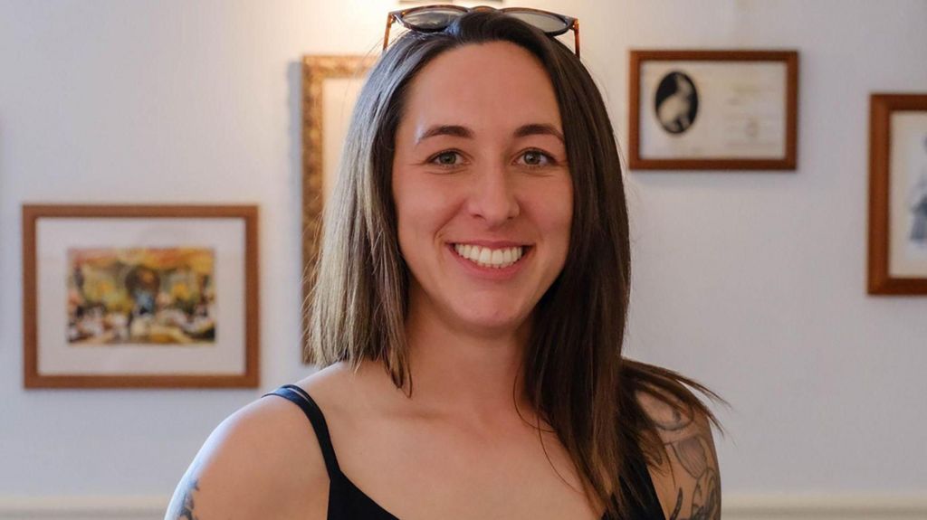 Amie Gray stands in a room with pictures on the wall. She has long, straight dark brown hair. Her black vest top shows tattoos running down both arms. She is smiling and has sunglasses on top of her hea
