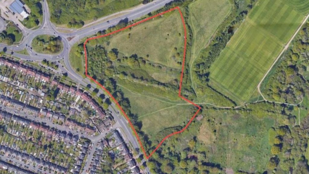 An aerial view of the proposed site, showing green fields to the right of a road and existing housing