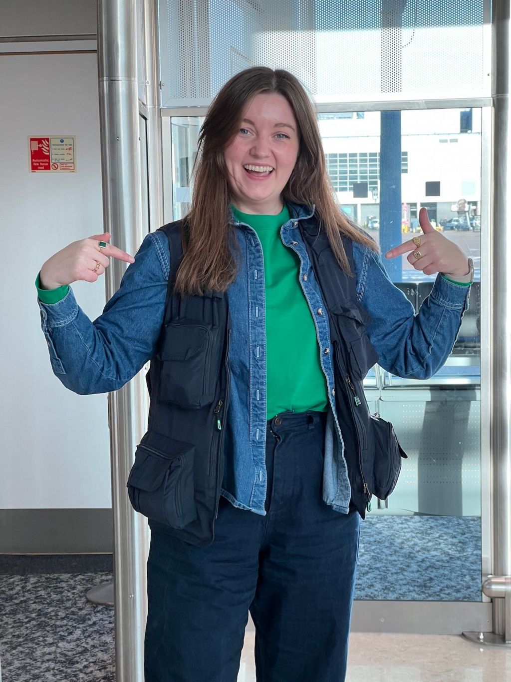 A young woman with long brown hair is in an airport departure lounge dressed in blue trousers and a denim shirt with a black fishing vest over the top. She is smiling and pointing both hands at her fishing vest.