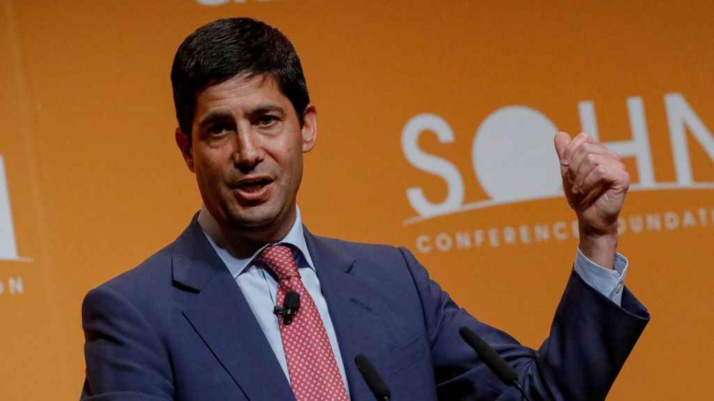 Kevin Warsh wearing a blue suit, light blue shirt and red patterned tie speaking at a conference in front of an orange background