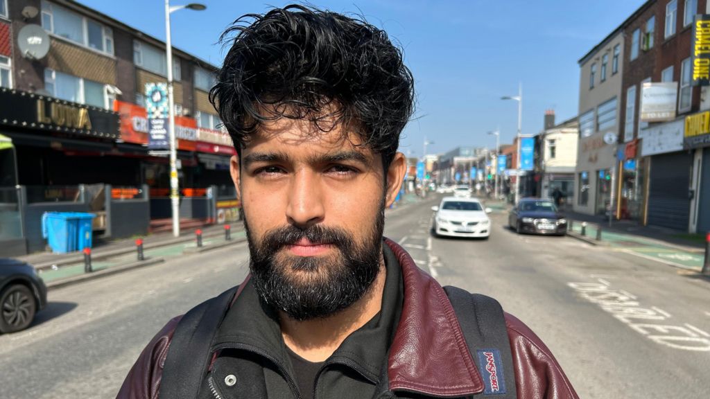 Sufiyaan Salam standing in the middle of Wilmslow Rd, Manchester, on the Curry Mile, with restaurants and shops on either side