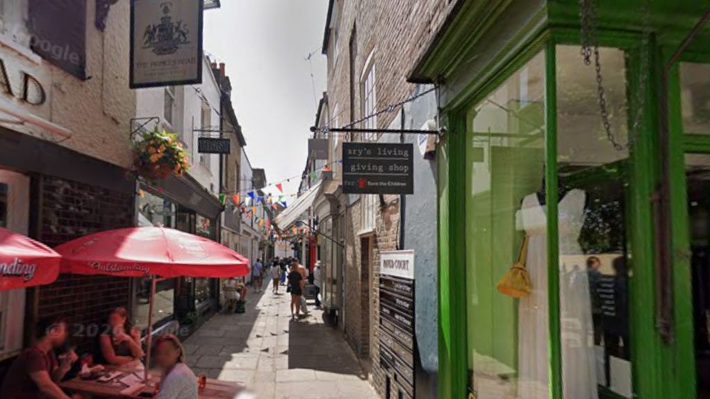 Paved Court, a narrow cobblestone pedestrian street. To the bottom left, people are sat having drinks below a red umbrella. Bunting goes across the street from  the shops' upper levels.