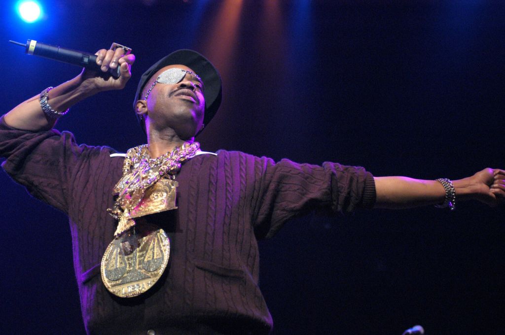 Slick Rick on stage. He is wearing a black baseball cap and a diamond-studded eyepatch, along with a plethora of oversized gold medallions