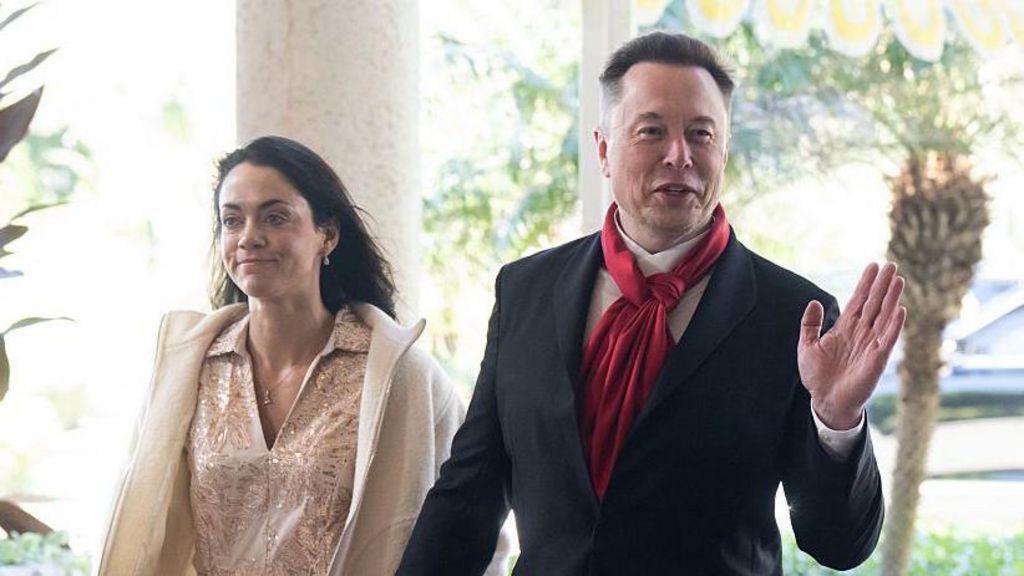 Shivon Zilis wearing a pale dress and jacket, with Elon Musk wearing a suit and red scarf. Both are smiling and Musk is waving.