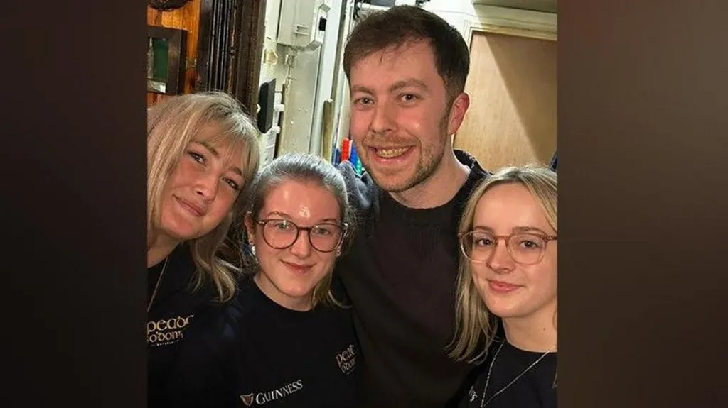 a smiling man stands in the middle of three women, each are wearing a black tshirt. they are in a pub. The man has short brown hair . the three women each have blond hair, two of them are wearing glasses.
