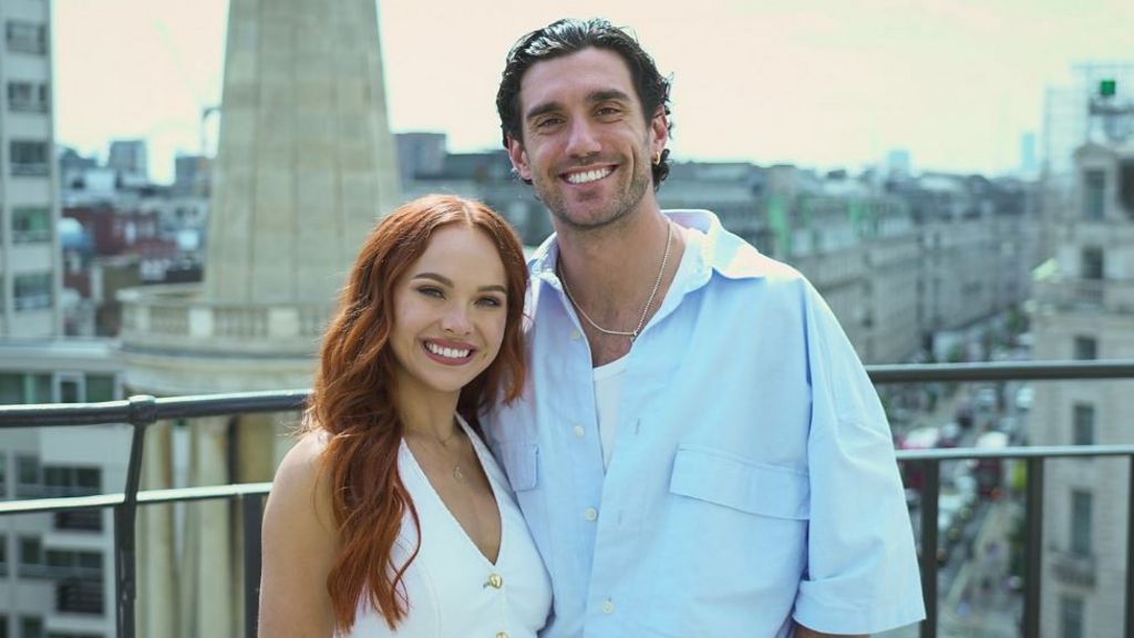 Alexis Warr, a woman in a white top with long, straight red hair, and Julian Caillon, a man in a blue shirt with dark hair slicked back, stand smiling on a balcony with a view of a street with cars and buildings