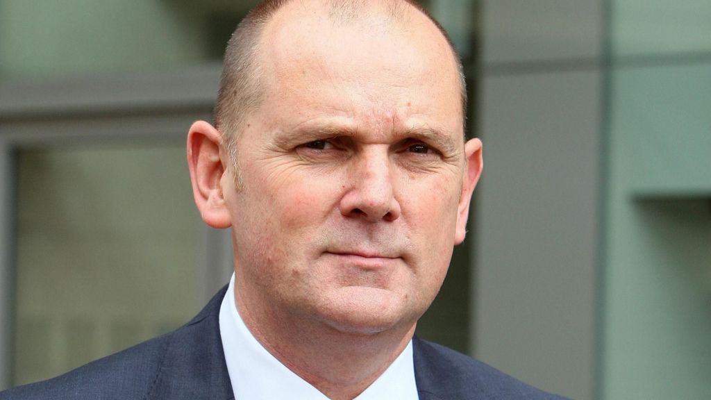 Head-and-shoulders image of Jim Gamble in a suit, looking directly at the camera with a neutral expression, taken outside a modern building.