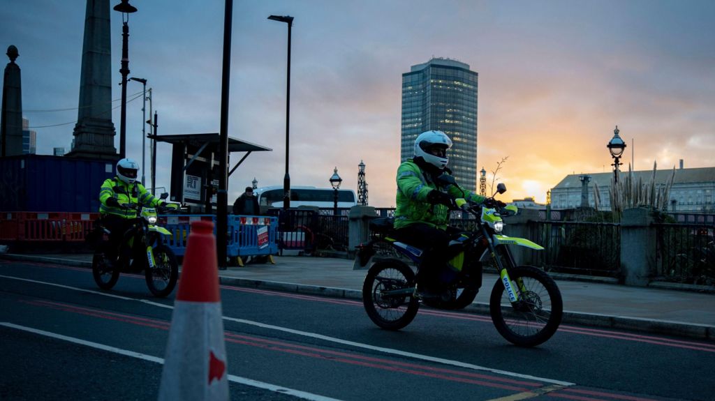 Two police officers on electric motorcycles patrol a London road at twilight. They wear white helmets and high-visibility jackets, with city landmarks and a sunset sky in the background.