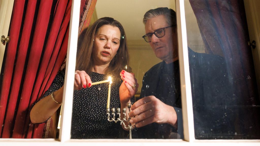 Victoria and Keir Starmer light first candle on menorah in windowsill. The picture is taken from someone outside, looking in to the room.
