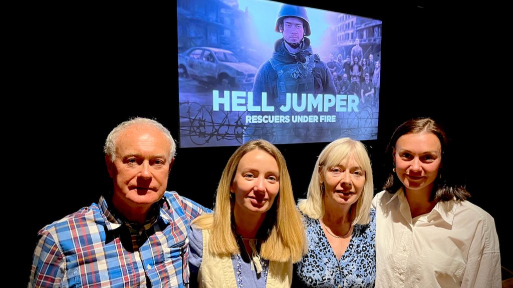 Rob Parry, Chris's dad, Chris's girlfriend Ohla Volodymyrivna Khomenko, mum, Christine Parry and Chris's sister, Kate Parry, standing in a row in front of a cinema screen showing a picture of Chris wearing helmet and protective clothing, withe wrds "Hell Jumper rescuers under fire" below.