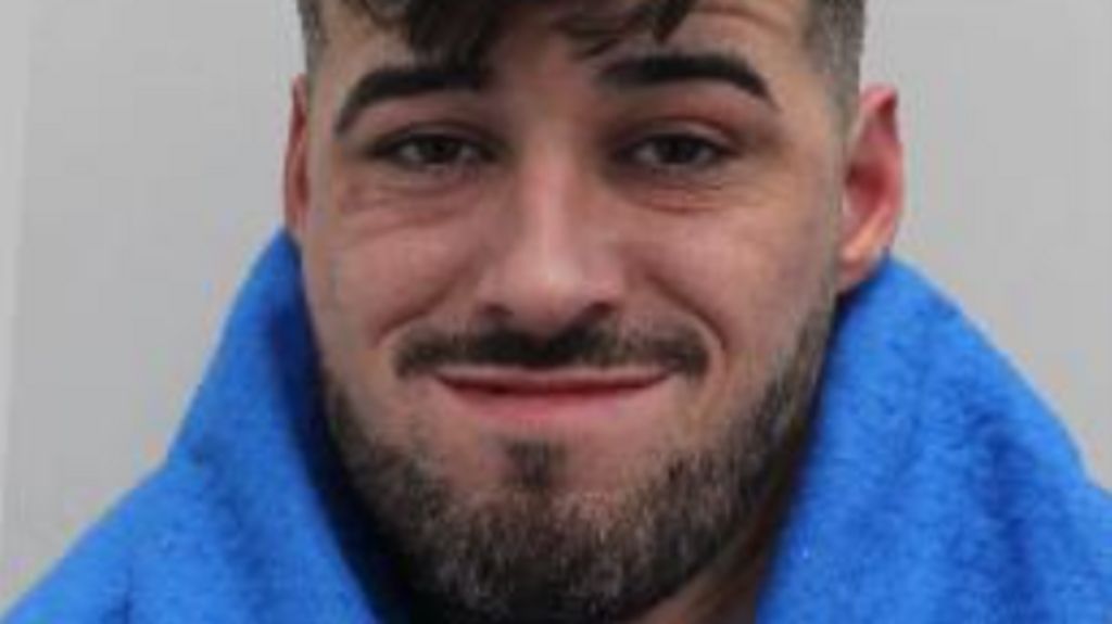 A close-up portrait of a man with short dark hair and a trimmed beard, wearing a blue garment and looking at the camera with a slight smile.
