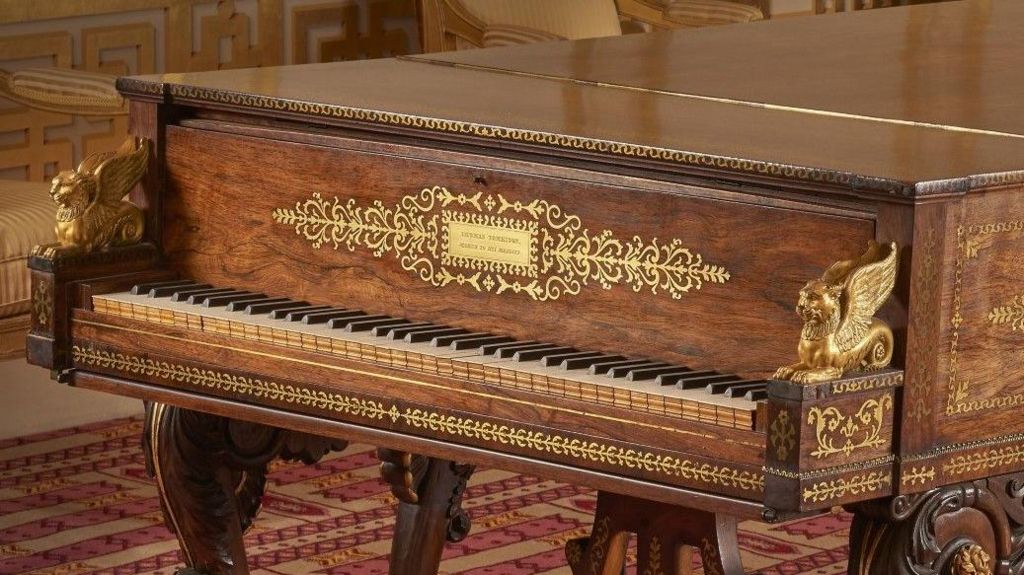 A close-up of gold and rosewood piano. It is elaborately decorated is in a room with a red and yellow carpet