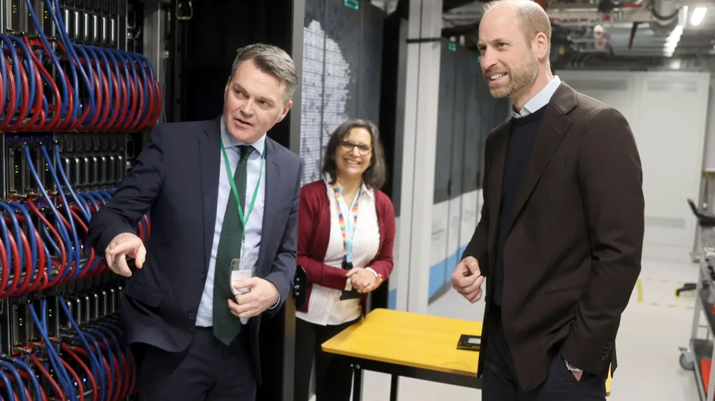 Prince William looks on as a man in a suit points out elements of the Isambard super computer. There are racks of blue and red cables, all looped around neatly and plugged in. A woman wearing a lanyard looks on cheerfully. 