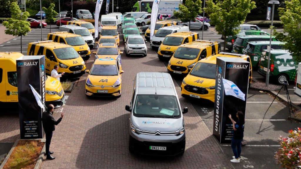 Electric vehicles including many commercially-branded vans are gathered in a car park, preparing to pass between two stands marking the start line of the EV Rally