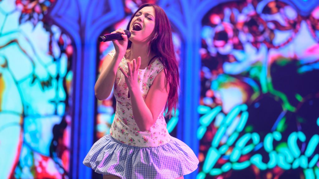 Alessi Rose is singing in to a microphone. She is on a stage which has a stainglass affect background. She wears a mini-dress which has a floral print top and a blue and white gingham-style skirt.
