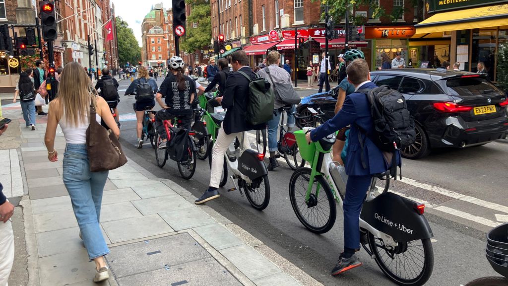 Cyclists, including Lime Bike users, wait at a red traffic light as pedestrians walk past