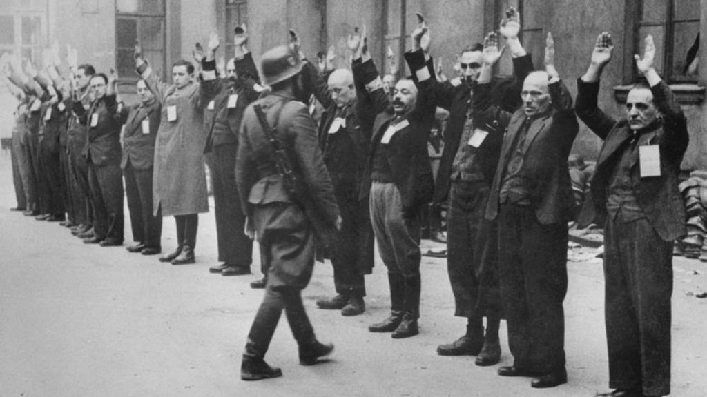 Judeus enfilerados e de mãos para cima são observados por um guarda nazista