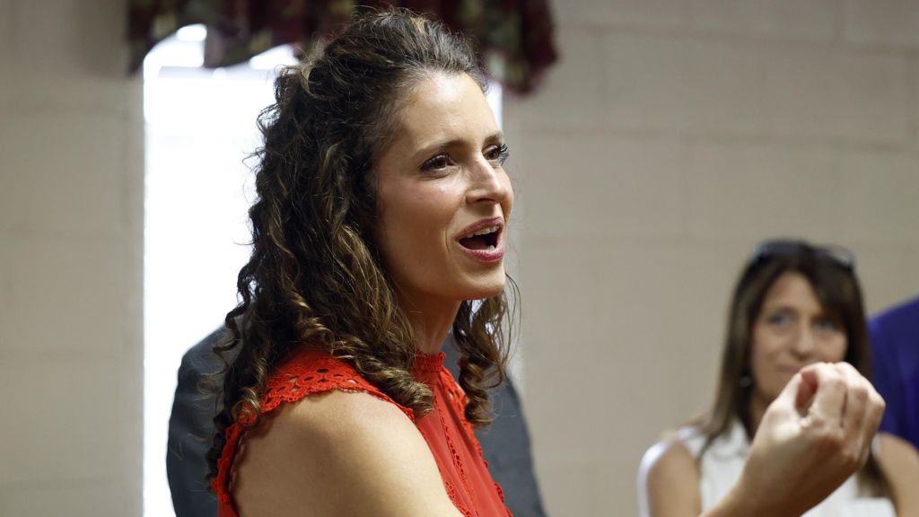 Julia Letlow is seen talking to constituents which smiling and appearing to talk. She is turned to the side and is seen from the shoulder up. She is wearing a red top.