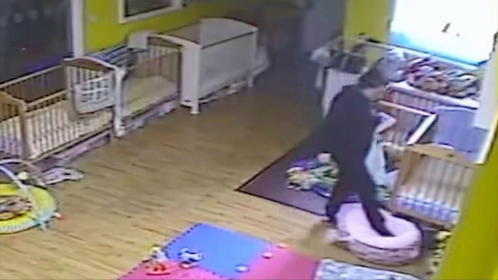 Image taken from a CCTV camera of a room at the nursery which has cots round the edges. There's a wooden floor and some mats. The walls are painted yellow. A woman, Kate Roughley can be seen on the right hand side, dressed in black. Her foot is on an empty bean bag - she is leaning forward as if moving it into position.