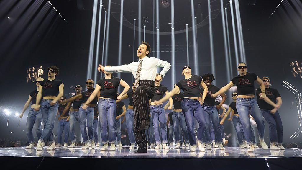 Harry Styles dances on stage at the Brit Awards. Wearing a shirt, tie and pinstripe trouser combination, he leads a group of dancers dressed in jeans and black t-shirts.