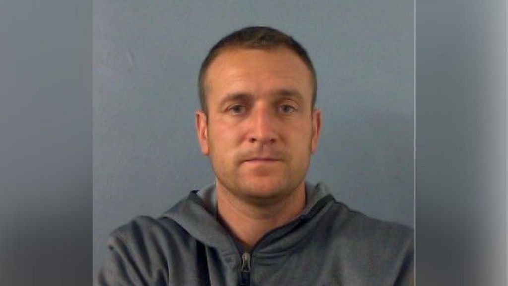 Police mugshot of James Sheen. He has short dark hair and is wearing a grey zipped hoodie
