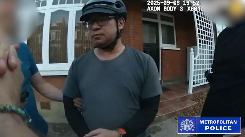 A still from bodycam footage of the police arrest of Vincent Chan. He is outside a house and is weaing a grey T-shirt with a black long-sleeved top underneath. He is wearing glasses and a bicycle helmet. Police are standing either side of him and his arm is being held.