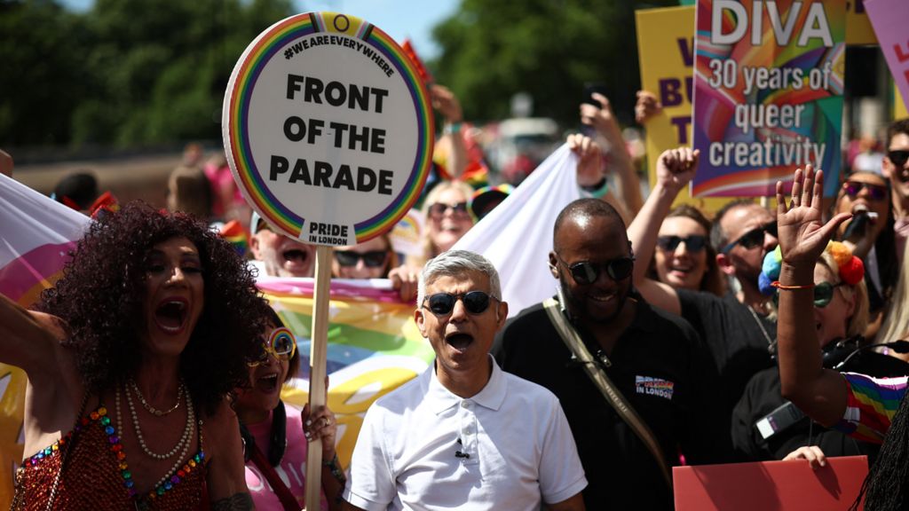Sadiq Khan, wearing sunglasses and a white polo top, cheers as he marches alongside other people cheering and waving. Some of them have banners, including one that reads "Diva, 30 years of queer creativity"