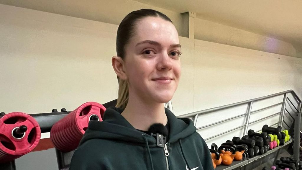 Megan has long brown hair tied up in a ponytail. She is wearing a dark hooded sweatshirt and is standing in a gym, positioned in front of shelves filled with colourful kettlebells, weight plates, and stacked dumbells.