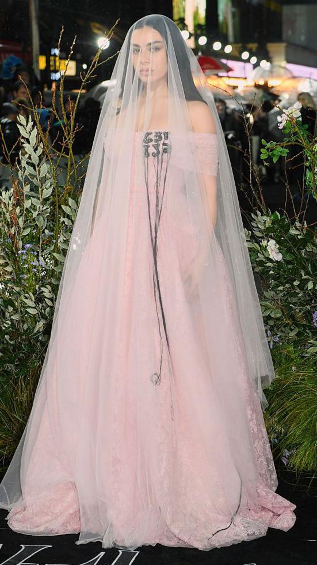 Charli XCX stands posing for a picture on the red carpet wearing a light pink floor-length dress with matching sheer veil that reaches the floor.