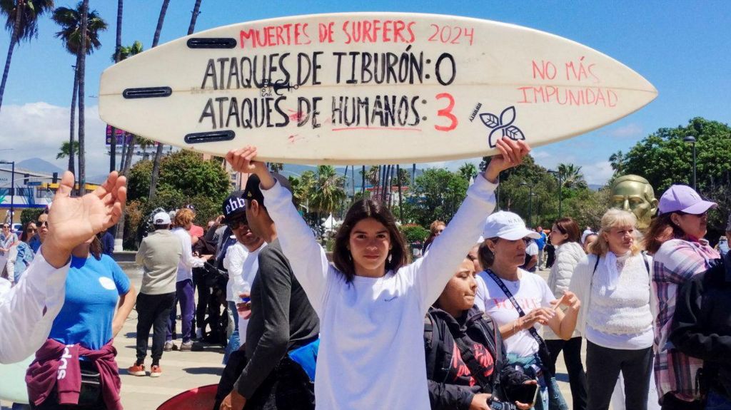 A woman holding a surf board with writing on it that says: 'Surfer deaths-Shark attacks: 0 and Human attacks: 3'