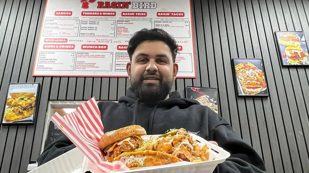 Wasim has short black hair and thick beard. He is wearing a black hoodie and is holding an open takeaway box filled with a fried chicken sandwich and two loaded tacos covered in sauce and shredded toppings. Behind him is a black wall with with vertical panels and colourful food posters. A large menu board labeled ‘Ragin’ Bird’ hangs above.
