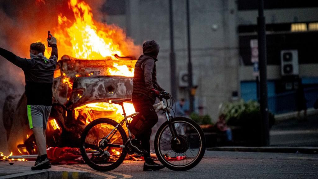 Two men covering their face celebrate, one holding a bottle of alcohol, while staring at the flaming wreckage of what was once a police car, now a fireball