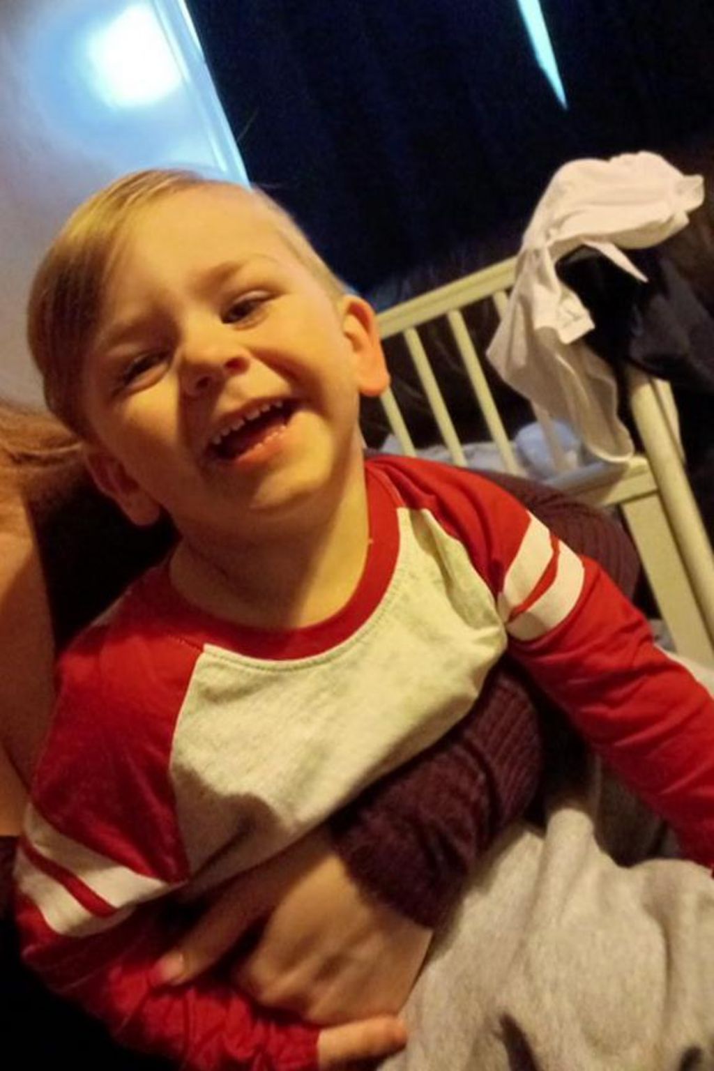 Kol Page, a toddler, grins at the camera. He has blond hair and is wearing a long sleeved T-shirt and is sitting on someone's lap (their face is not visible) in front of a cot.