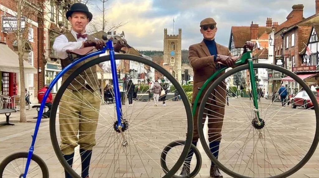 Two men standing in the middle of a pedestrianised high street with their penny farthings