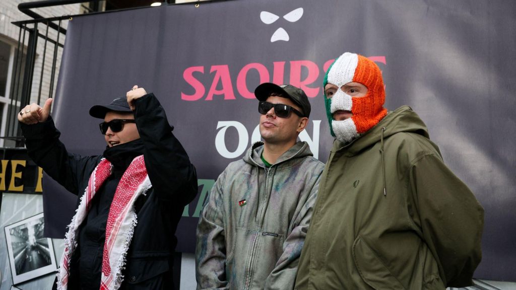 Mo Chara, Mógalí Bap and DJ Próvaí stand in front of a black screen with the band's logo and Saoirse don Phalaistín (Irish for Freedom for Palestine) written in the Palestinian flag colours. Mo Chara is on the left wearing dark clothing and a red and white keffiyeh. Mógalí Bap is wearing sunglasses, a cap and a grey zip hoodie. Dj Próvaí is wearing an Irish tricolour balaclava and a green jacket.