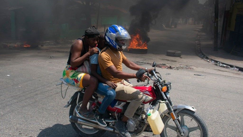 Three people ride a motorcycle along a street, with a burning tyre in the background