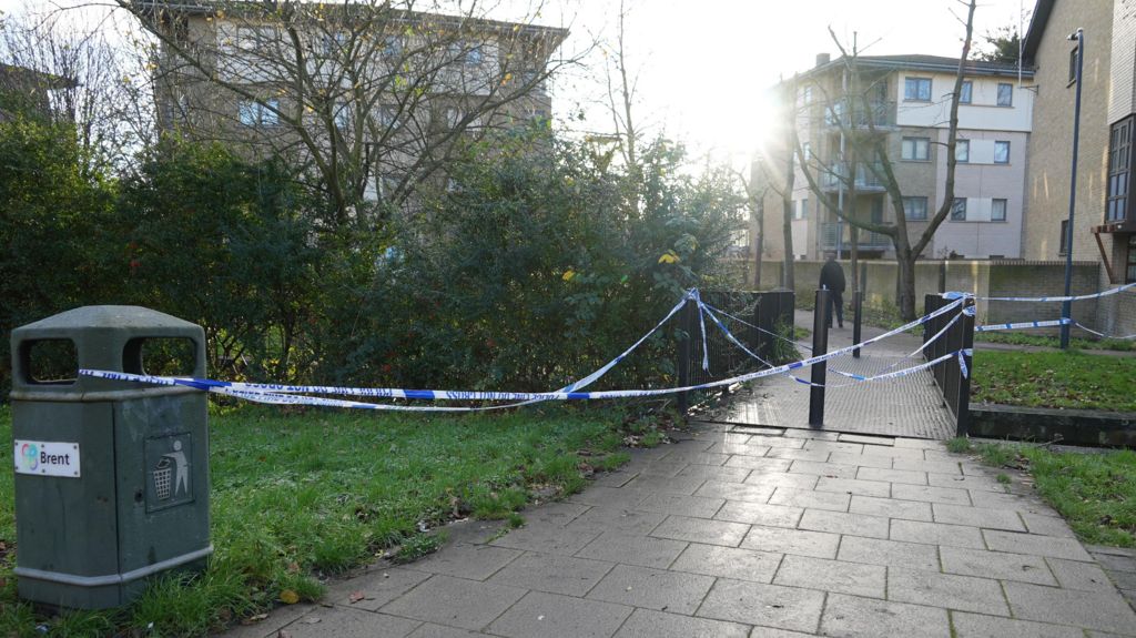 A police cordon over a pathway, attached to metal fencing, a lampost and a bin.