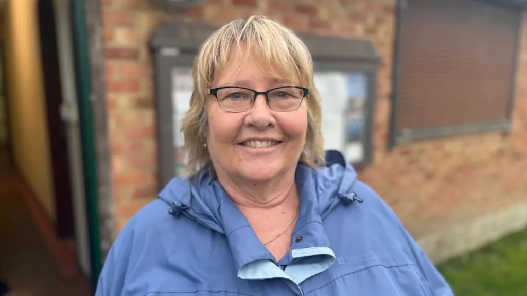 A woman pictured outside a building wearing reading glasses and a blue raincoat. She has short blonde hair and is smiling.