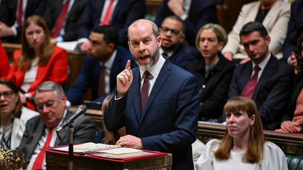 The Business Secretary Jonathan Reynolds addresses the House of Commons, with two rows full of MPs behind him. He has short grey facial hair and wears a dark blue suit with a white shirt and maroon tie.